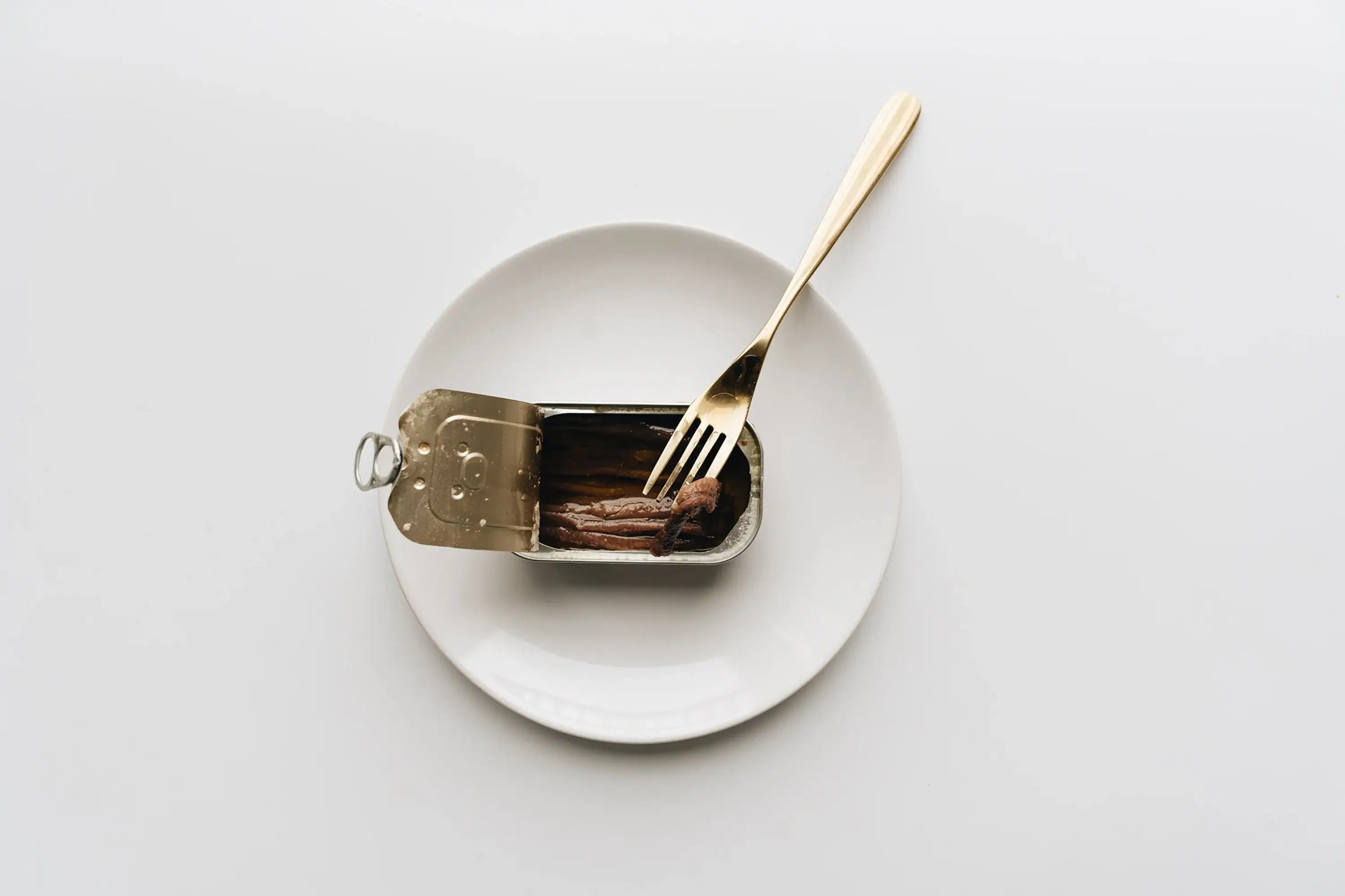 an open can of tinned fish on a white plate with  gold fork